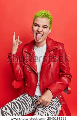 Sassy punk girl sticks out tongue shows heavy metal sign gesture feels cool has trendy green hairstyle wears leather jacket sits on chair isolated over red background enjoys rock music. Youth concept Royalty-Free Stock Photo #2040198566