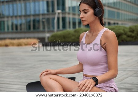 Photo of relaxed woman in sportsclothes sits crossed legs on fitness mat listens relaxing music practices yoga in downtown enjoys relaxation and meditation. Urban background. Healthy lifestyle