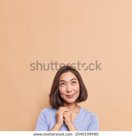 Indoor shot of good looking young woman with dark hair eastern appearance keeps hands together looks with hopeful dreamy expression upwards wears casual jumper isolated over beige background
