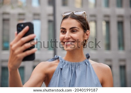 Beautiful stylish woman makes video call via smartphone while strolling in urban setting enjoys summer day has upbeat mood poses against blurred background. Pretty female model has online conversation