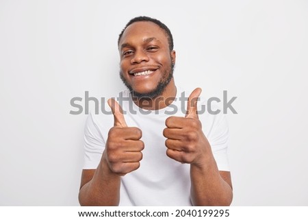 Happy smiling dark skinned man approves incredible promo keeps thumbs up likes and agrees being satisfied with something wears casual clothes praises cool job isolated over white background.