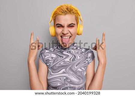 Sassy hipster girl with trendy yellow hair vivid makeup shows heavy metal horns gesture sticks out tongue has fun listens music during free time partying at concert enjoys event likes smth cool