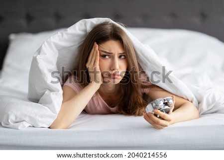 Unhappy young Caucasian woman lying in bed with alarm clock, not willing to get up in morning. Upset millennial lady having problem waking up after sleepless night, cuddling under warm blanket Royalty-Free Stock Photo #2040214556