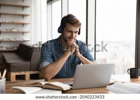 Smiling young Caucasian male student in earphones use computer study online from home. Happy millennial man wear headphones have online video lesson or webcam course or training on laptop. Royalty-Free Stock Photo #2040223433