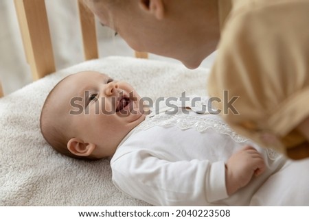 New mom talking to happy adorable few month baby resting in crib and smiling. Young mother speaking and playing with infant son or daughter lying on back. Child care, motherhood concept Royalty-Free Stock Photo #2040223508