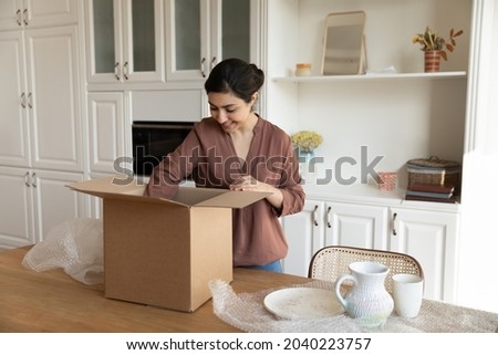 Just after relocation. Happy biracial woman unpack stuff take ceramic tableware out of big carton container at modern kitchen. Young indian lady get internet purchase open box with newly bought dishes Royalty-Free Stock Photo #2040223757