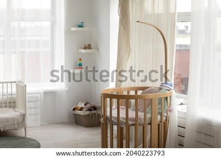 Kids bedroom with wooden bassinet for newborn infant at window, white crib and walls, toys, pale decoration colors. Baby room interior with nobody. Furniture for nursery, home for child concept