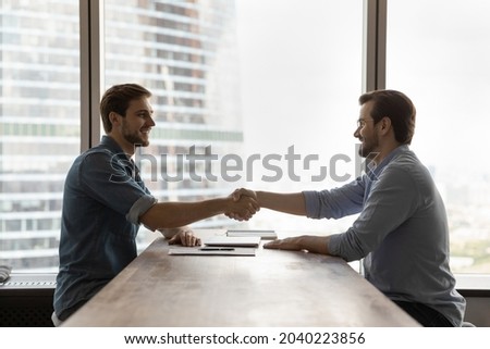 Smiling young Caucasian businessmen handshake get acquainted greeting at meeting in office. Happy male employees colleagues shake hands close deal make agreement after successful negotiation. Royalty-Free Stock Photo #2040223856