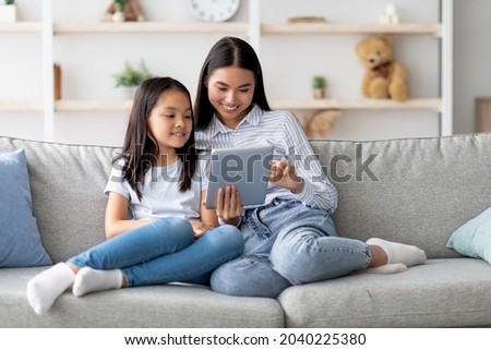 Happy young asian mom and daughter using digital tablet, watching videos or surfing internet, sitting on sofa at home, copy space. Young korean family using pad together