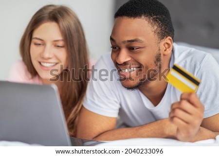 Portrait of happy multiracial couple with credit card and laptop lying on bed, shopping online from home. Millennial black man and his wife purchasing on sale in web store, using contactless payment