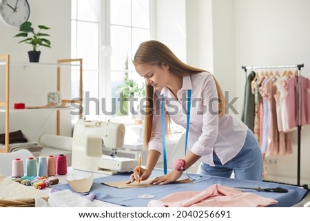Seamstress at work. Dressmaker making clothes in modern studio. Tailor holding pencil and marking fabric. Woman standing at table with cut textile, sewing machine, thread, pins, needles, tape, cutouts Royalty-Free Stock Photo #2040256691