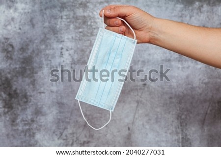 A man's right hand holding the rubber band of a blue surgical mask on a textured gray background.