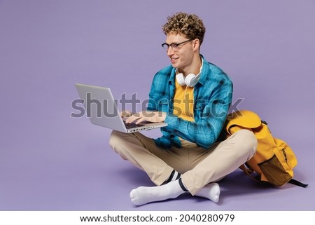 Full body young boy teen student in casual clothes backpack headphones glasses sit hold work on laptop computer isolated on violet background studio Education in high school university college concept Royalty-Free Stock Photo #2040280979