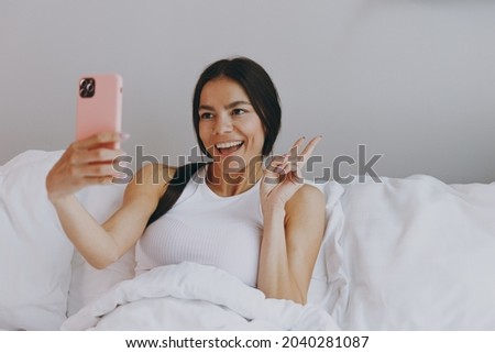 Excited young woman in white tank top sit in bed doing selfie shot on mobile cell phone post photo on social network in bedroom lounge home in own room house wake up dream be lost in reverie good day