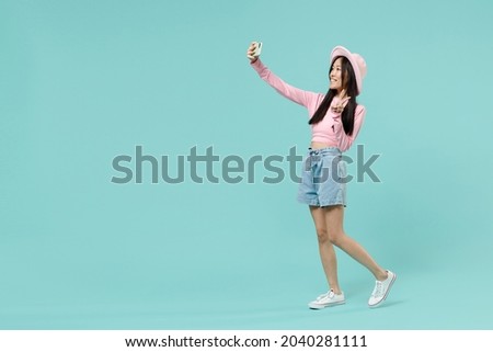 Full size body length fun young brunette asian woman 20s wears pink clothes doing selfie shot on mobile phone showing victory sign strolling isolated on pastel blue color background studio portrait.