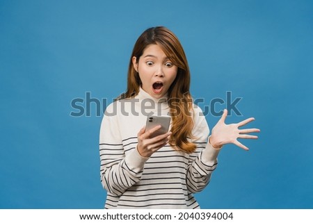 Surprised young Asia lady using mobile phone with positive expression, smiles broadly, dressed in casual clothing and standing isolated on blue background. Happy adorable glad woman rejoices success. Royalty-Free Stock Photo #2040394004