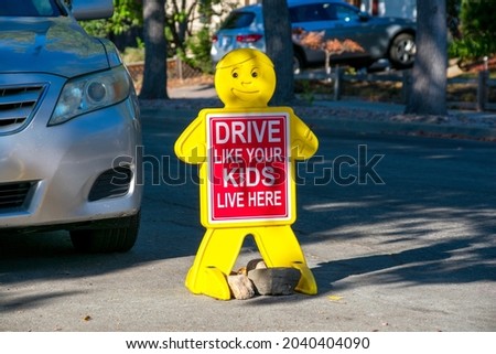 Drive Like Your Kids Live Here road sign in residential neighborhood near parked car. Child like yellow figure with red safety sign on road.
