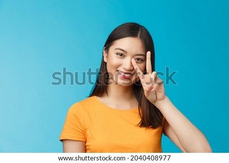 Positive cute and silly asian girlfriend posing photograph lovely smiling, show peace victory sign near face grinning friendly, carefree enthusiastic mood, stand blue background in yellow t-shirt