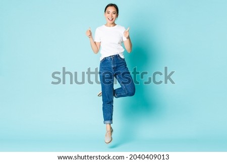 Fashion, beauty and lifestyle concept. Upbeat pretty asian girl in casual outfit, enjoying shopping, jumping from happiness and excitement, showing thumbs-up over light blue background