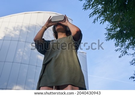 A girl holds virtual reality glasses with her hands, standing against the background of a mirrored building. The concept of the future. High quality photo