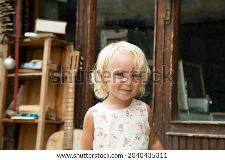 Little blond blue eye girl in white dress with pink flowers portrait. Tricky funny child. Retro style picture. Vintage door with bookshelf and guitar in background. Seppia. Childhood in old town 