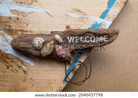 An object decorated with shells on a wooden board with paint by DIY and driftwood with "WELCOME" written on it.