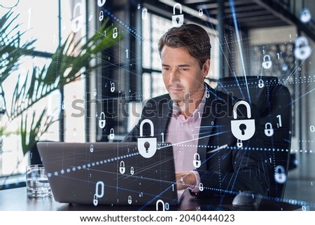 Handsome businessman in suit at workplace working with laptop to defend customer cyber security. Concept of clients information protection and brainstorm. Padlock hologram over office background.