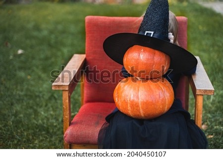 Scary little girl in witch costume, hat with many pumpkins celebrating halloween holiday. Sitting on armchair in coat with pumpkin. Stylish image. Horror, fun at children's party in barn on street.
