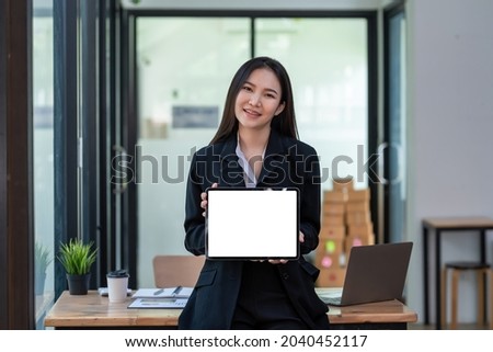 Front view of Asian woman standing holding blank white screen tablet at office. Looking at camera. Mock up.