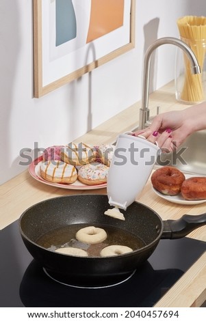 Shot of a female hand that holds a donut mold and pours a liquid dough in a black pan with hot oil. A white donut mold above a pan. Donuts, picture, jar of spaghetti and sink on the background.