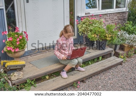 a girl child with a laptop is sitting on the porch of the house during a lesson on a remote photo without a filter