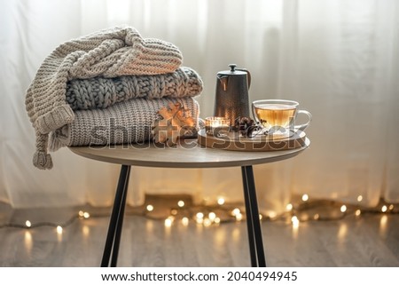 Home autumn composition with tea and knitted sweaters in the interior of the room, on a blurred background with a garland.