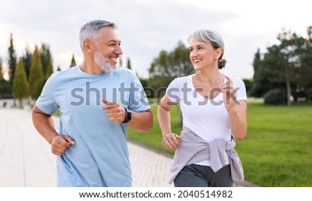Happy senior husband and wife in sportive outfits running outdoors in city park, lovely retirees couple jogging in sunny morning looking at each other with warmth and smile. Healthy lifestyle concept