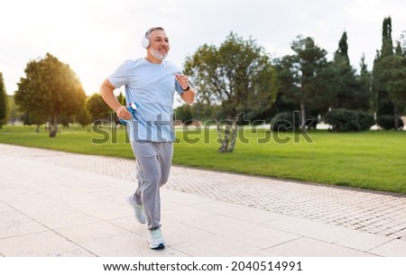 Full-length photo of happy senior man in headphones and sportswear jogging running outside in city park with water bottle in hand. Healthy active lifestyle on retirement and sport outdoor concept
