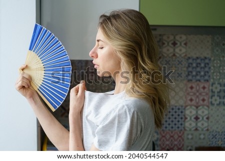 Side view caucasian tired overheated woman in t-shirt using wave fan suffer from heat sweating, cools herself, closed eyes feels sluggish due problem no air conditioner at home at summer weather. Royalty-Free Stock Photo #2040544547