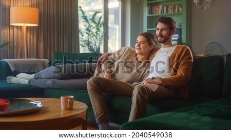 Couple Watches TV together while Sitting on a Couch in the Living Room. Girlfriend and Boyfriend embrace, cuddle, talk, smile and watch Television Streaming Services. Home with Cozy Stylish Interior. Royalty-Free Stock Photo #2040549038