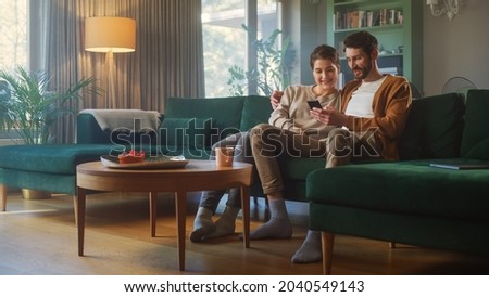 Couple Use Smartphone Device, while Sitting on a Couch in the Cozy Apartment. Boyfriend and Girlfriend Talk, do e-Shopping on Internet, Watching Funny Videos, Use Social Media, Streaming Service. Royalty-Free Stock Photo #2040549143