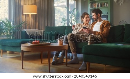 Couple Use Smartphone Device, while Sitting on a Couch in the Cozy Apartment. Boyfriend and Girlfriend Talk, do e-Shopping on Internet, Watching Funny Videos, Use Social Media, Streaming Service. Royalty-Free Stock Photo #2040549146