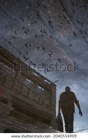 Blurred silhouette of reflection of one person walking alone on wet city sidewalk on rainy day. Birds are flying across the sky. Abstract photography.