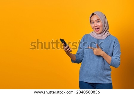 Portrait of excited attractive Asian woman pointing finger at mobile phone over yellow background
