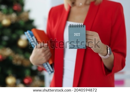 Businesswoman in red jacket holds calendar for 2022 against background of New Year tree