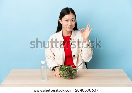 Young Chinese girl eating a salad showing ok sign with fingers