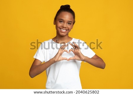 Happy teen african american girl pupil blogger in white t-shirt show heart gesture with hands, isolated on yellow background. Love sign, Valentine day, kids positive emotions and study, covid-19 Royalty-Free Stock Photo #2040625802