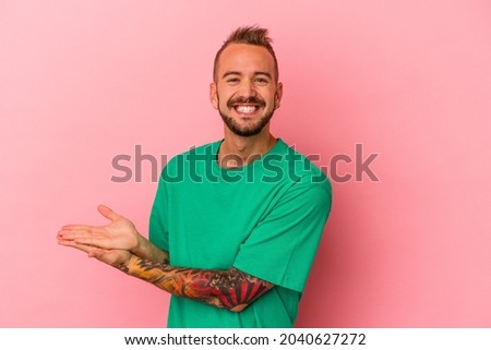 Young caucasian man with tattoos isolated on pink background  holding a copy space on a palm.