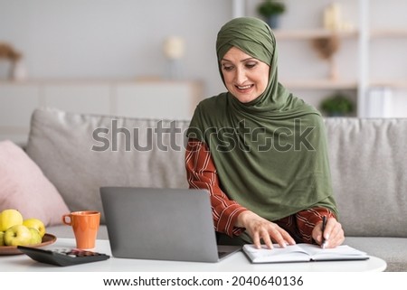 Senior Islamic Businesswoman Taking Notes Sitting At Laptop And Working Online At Home. Muslim Lady Making Financial Report Indoor. Female Business Career And Entrepreneurship, Remote Job Concept