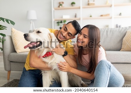 Portrait of happy multiracial couple scratching their pet dog, sitting on floor at home. Arab guy and his Caucasian girlfriend hugging their golden retriever in living room Royalty-Free Stock Photo #2040640154