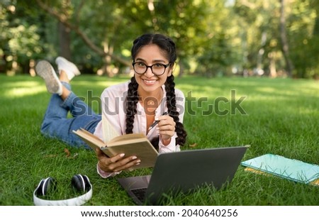 Studentship. Indian student girl learning with laptop computer and book, taking notes, lying in park outdoors, free space. University education, college lifestyle and e-learning Royalty-Free Stock Photo #2040640256