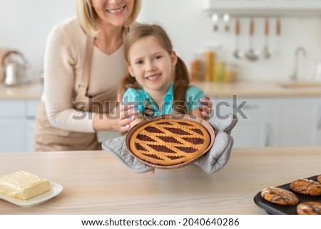 Family Recipe, Cooking Food With Love. Happy little girl holding and showing freshly baked pie close up to camera, preparing homemade pastry with granny in kitchen, adorable female child wearing apron Royalty-Free Stock Photo #2040640286