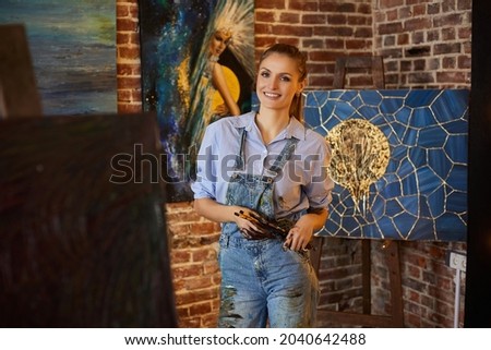 Portrait of young smiling female artist with her artworks in art studio. Painter holding art brushes. Creative process, relaxation, leisure, hobby, stress management.