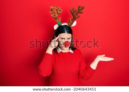 Young hispanic woman wearing deer christmas hat and red nose confused and annoyed with open palm showing copy space and pointing finger to forehead. think about it. 